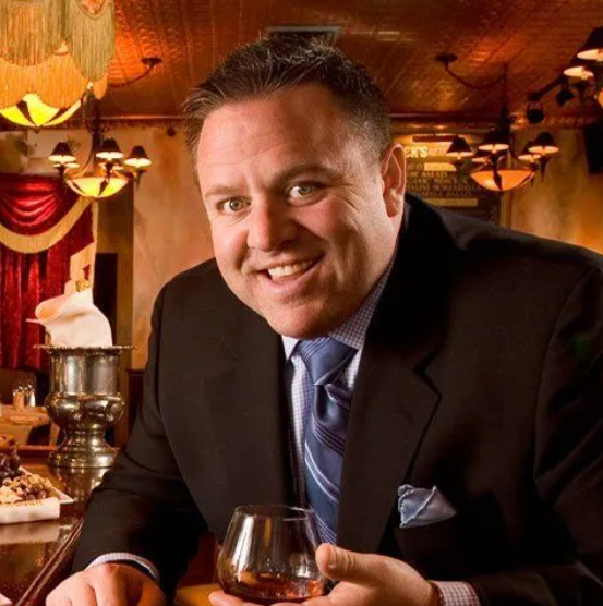 willie degel of uncle jacks in a suit and tie is holding a glass of whiskey .