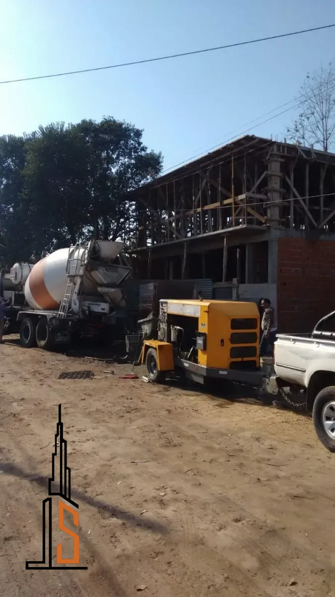 Camión de cemento vertiendo hormigón en una obra. Maquinaria amarilla, edificio a medio construir, camino de tierra marrón.
