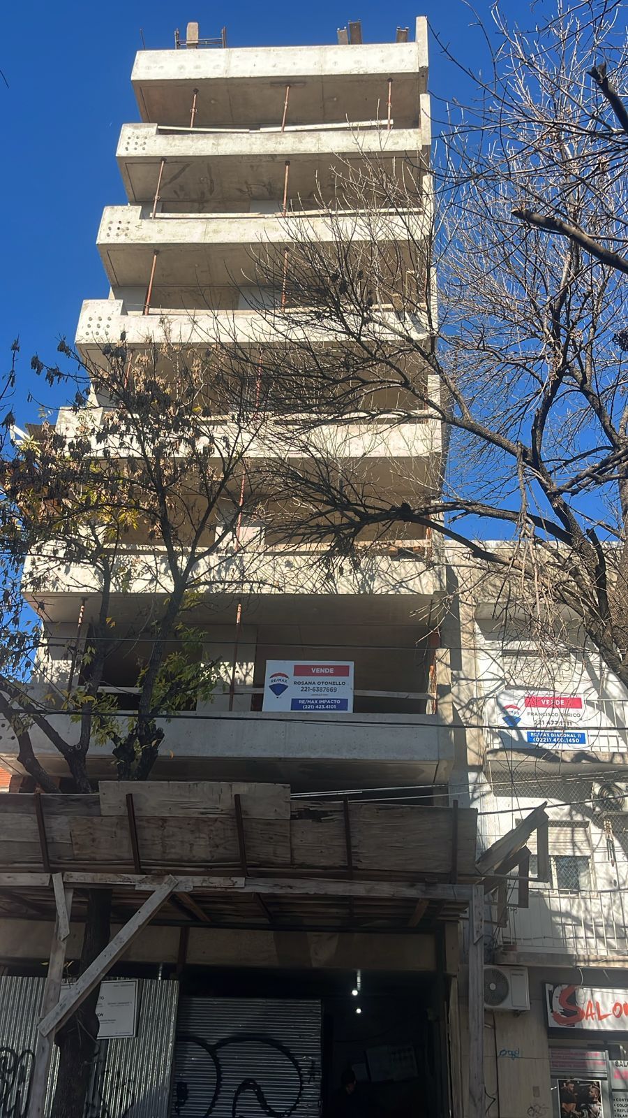 Un edificio alto de hormigón de varias plantas con balcones a la vista. Hay un árbol y un letrero en la entrada.