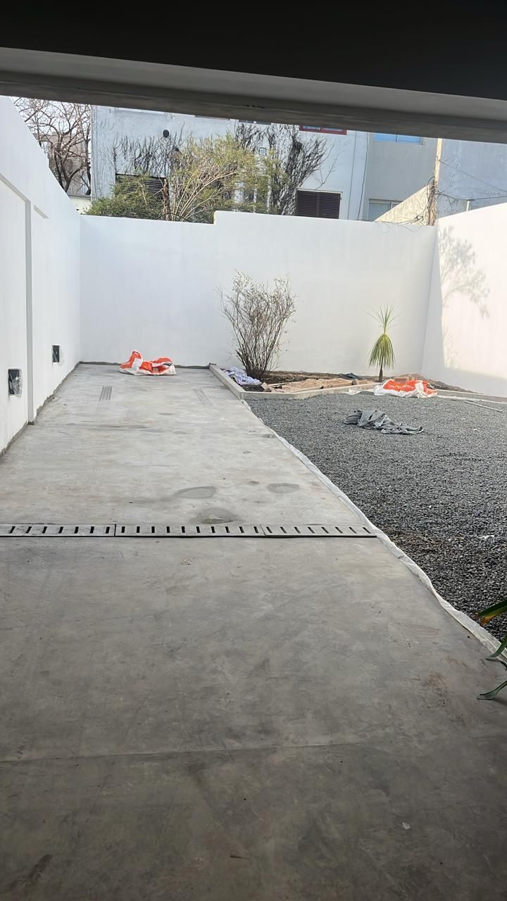 Patio de hormigón con grava y paredes blancas. Plantas pequeñas y un arbusto en la zona de grava.