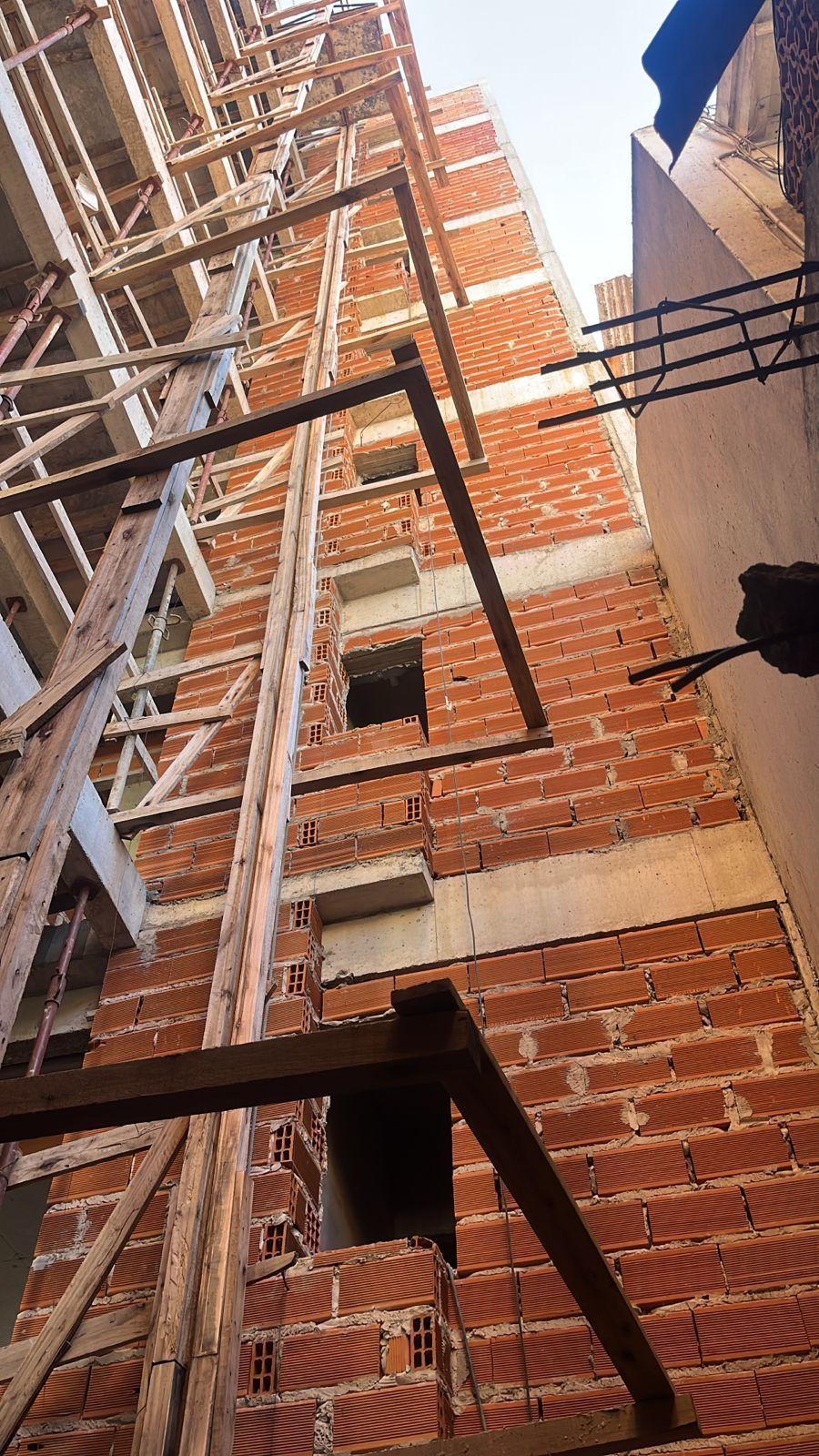 Edificio de ladrillo en construcción, al que se fijan andamios de madera y vigas metálicas en la fachada.