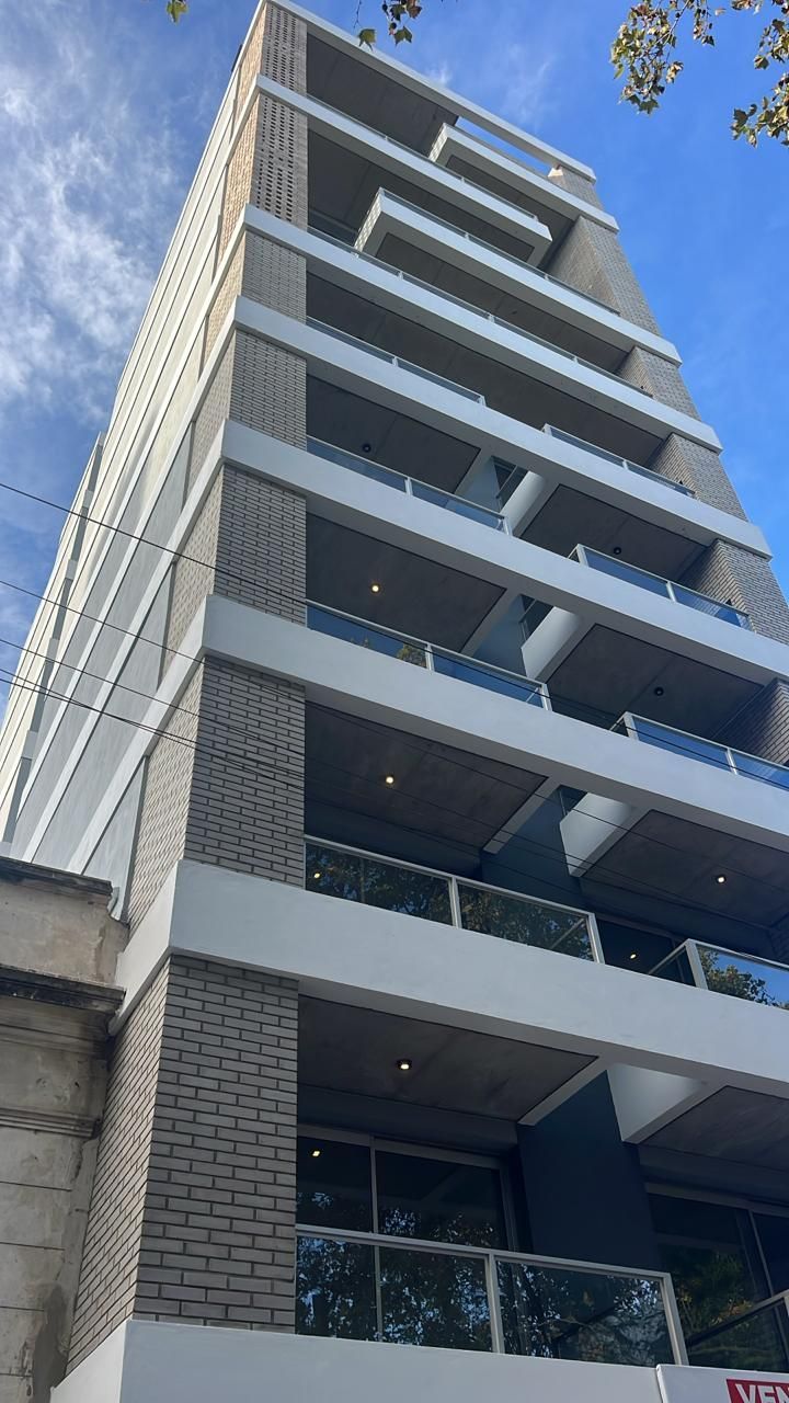 Edificio de apartamentos alto y moderno con balcones, fachada gris y blanca, contra un cielo azul.