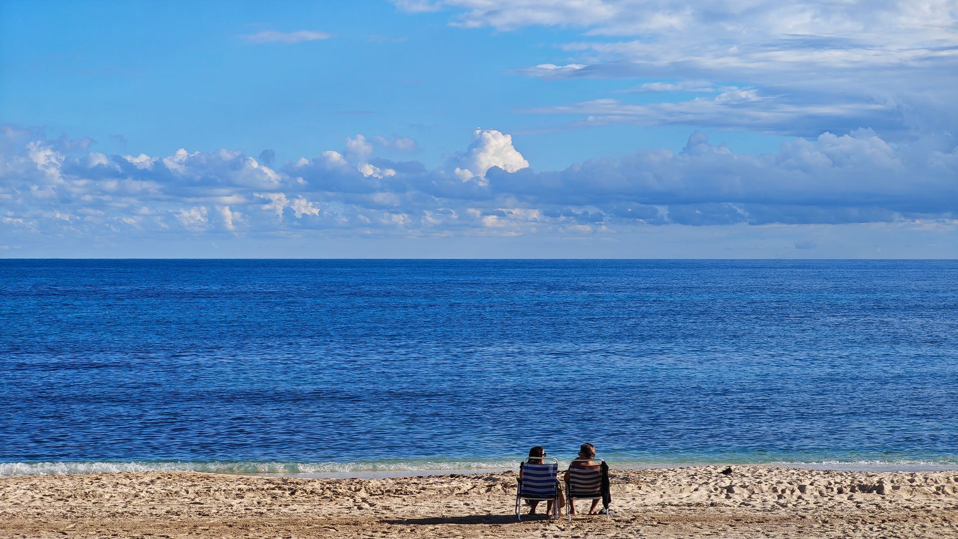 Two figures on beach, facing vast blue ocean under partly cloudy sky.