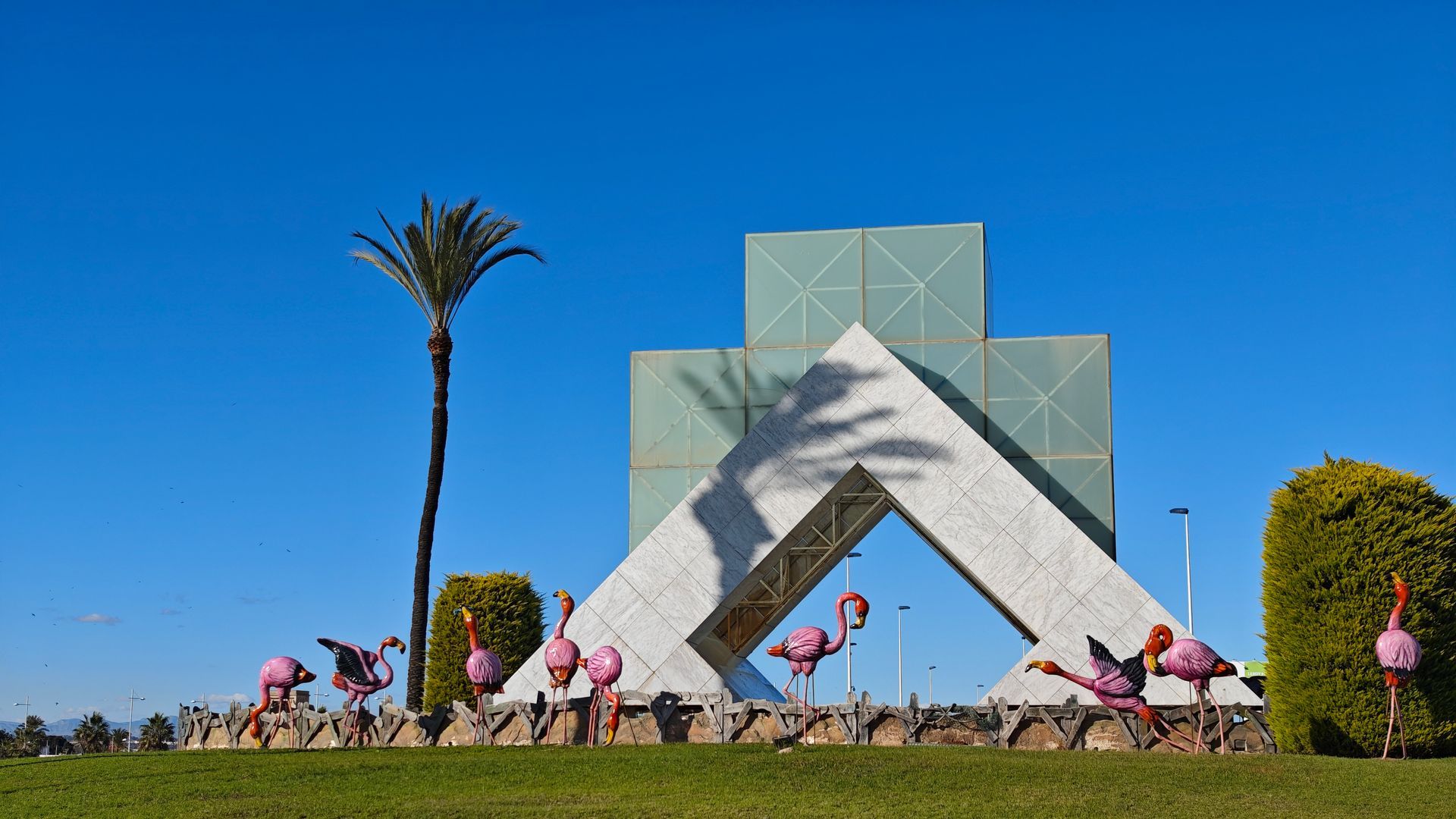 A monument with a geometric design and pink flamingo statues. Bright blue sky.