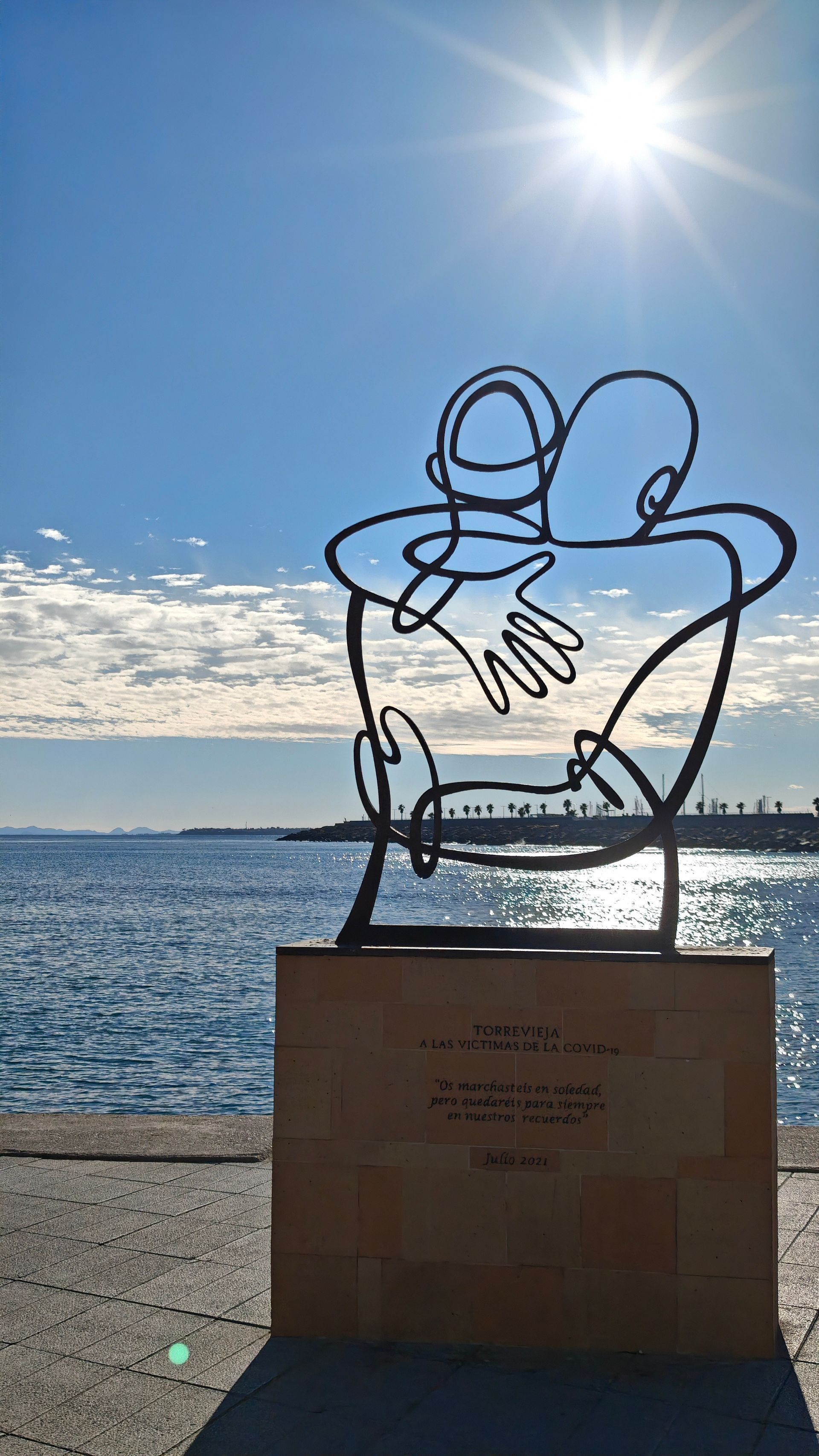 Metal sculpture of two embracing figures, with ocean and sunlit sky in background.