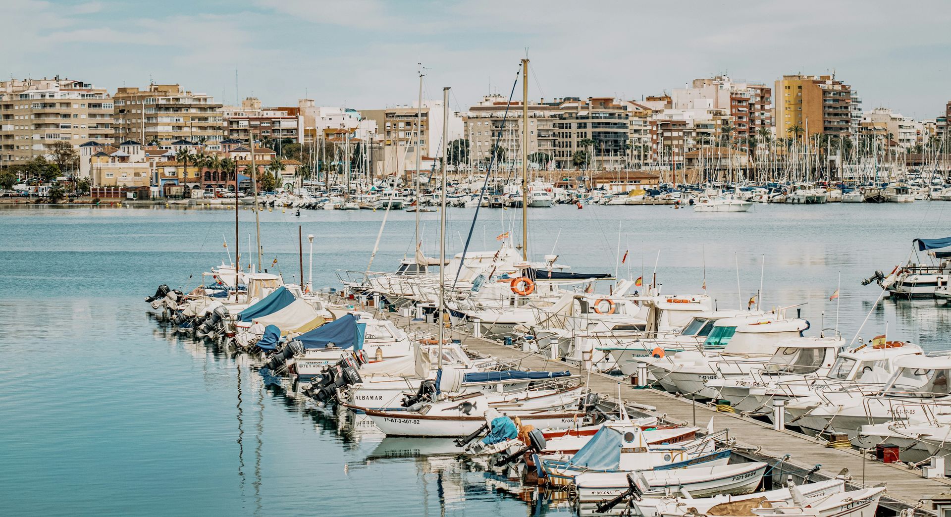 Boats docked in a harbor, city buildings in the background. Bright day, blue water.