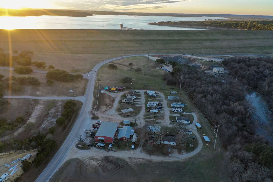 Aerial view of a small campground by water at sunset, with parked vehicles and tents amid trees and dirt roads