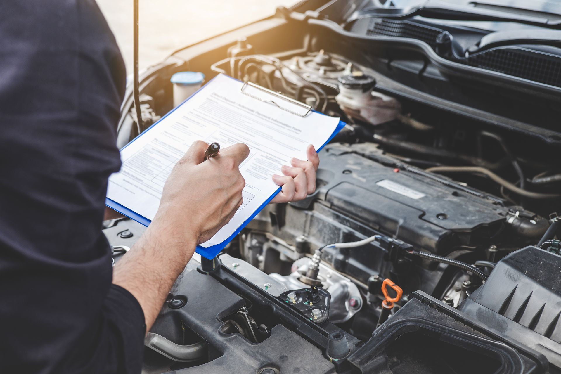 A man is looking under the hood of a car while holding a clipboard.