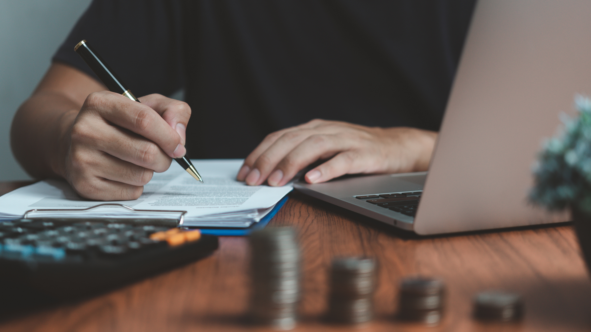 person holding a clipboard with papers about to sign. calculator, change, and a laptop are around him.