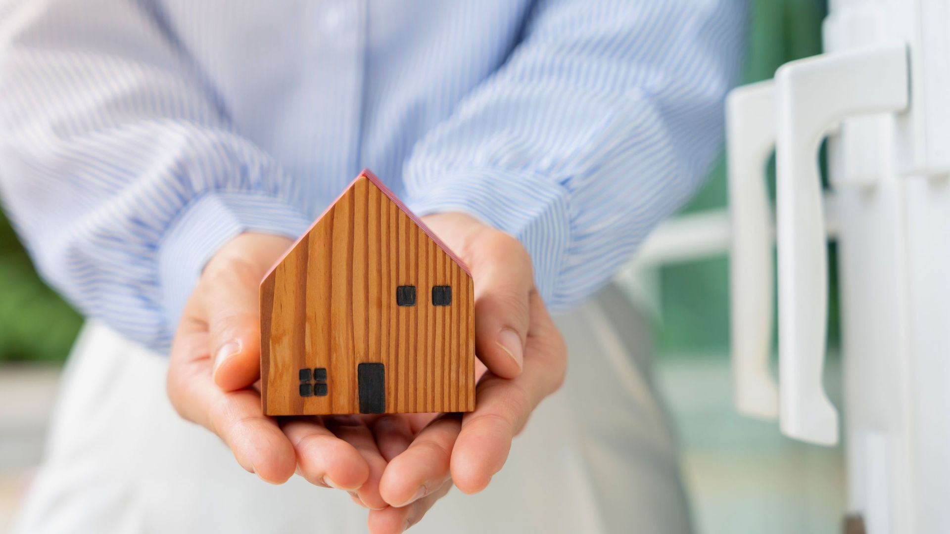 person wearing a blue and white button down shirt holding a small wooden house with both hands