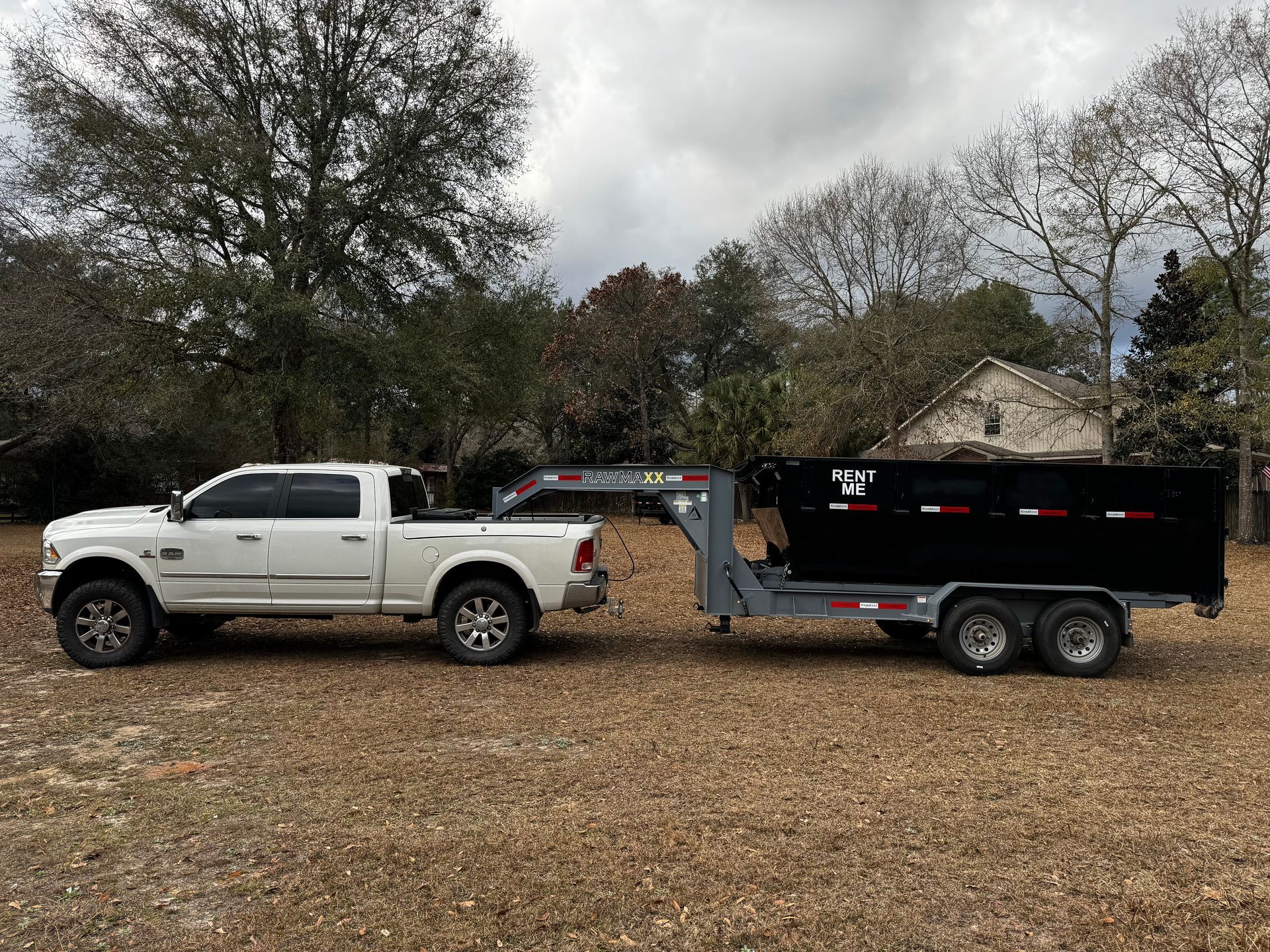 White truck towing a black dump trailer on a grassy field; trees and house in the background.