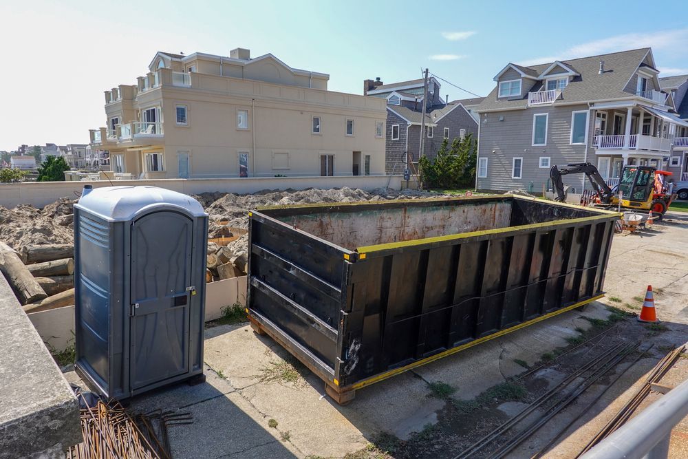 Construction site with a portable toilet and a large dumpster. Buildings in the background.
