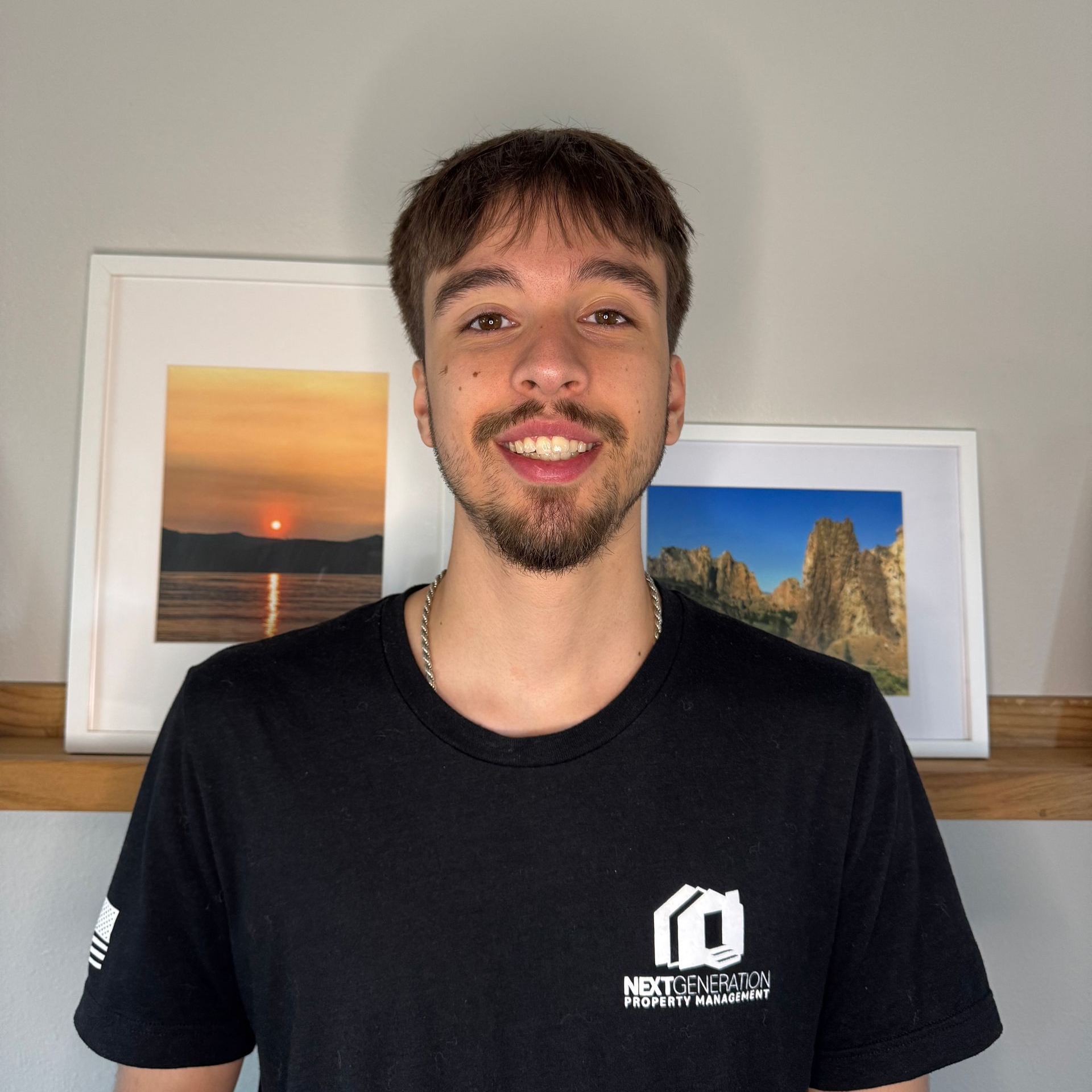 Smiling man with brown hair, goatee, and black shirt. Background features artwork and a blurry wall.