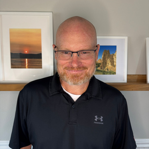 A bald man with a beard and glasses is wearing a black polo shirt.