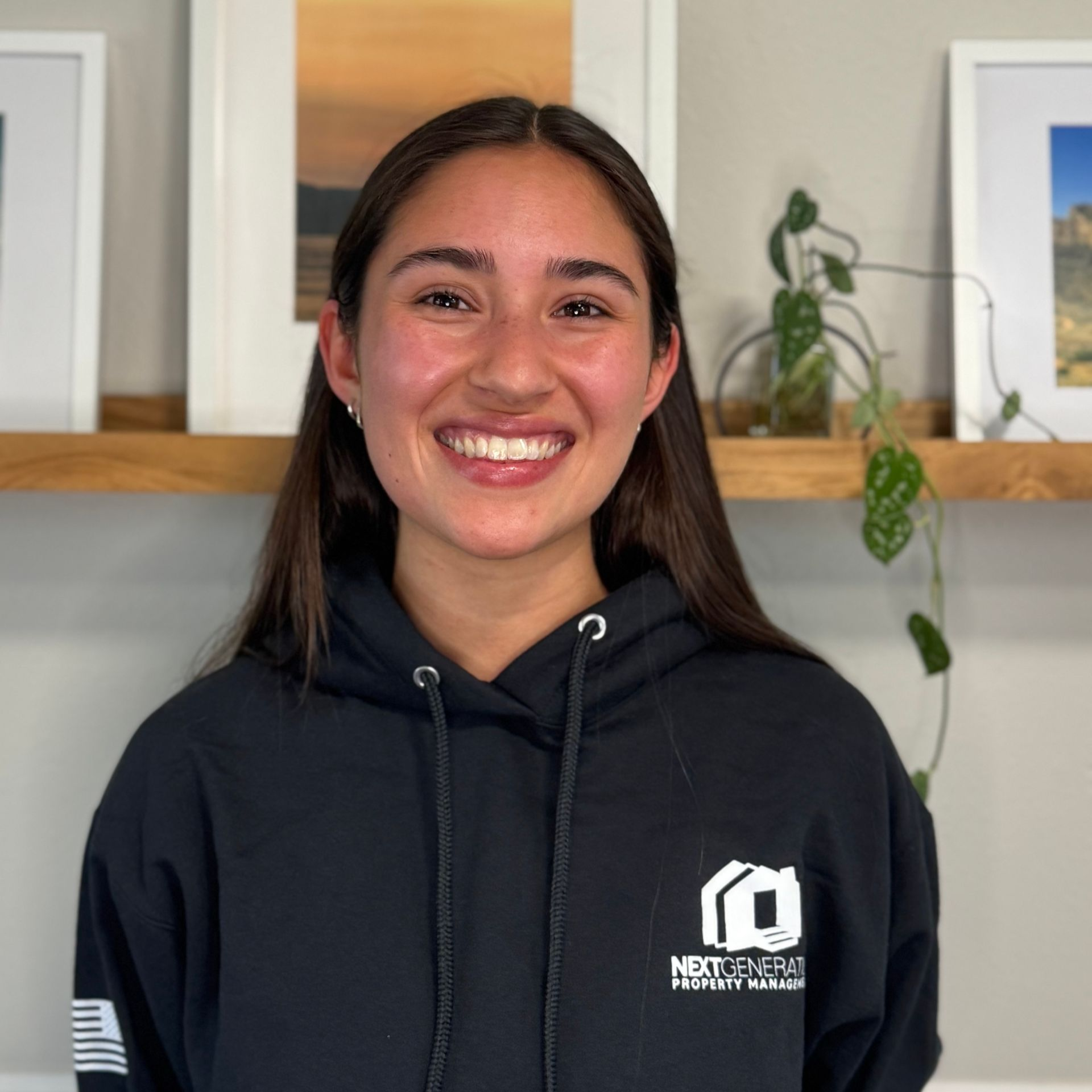Woman smiling, wearing a black hoodie with logo, standing in front of framed artwork and a shelf with plants.
