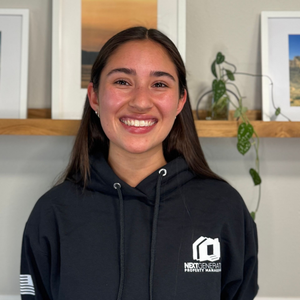 Woman smiling, wearing a black hoodie with logo, standing in front of framed artwork and a shelf with plants.