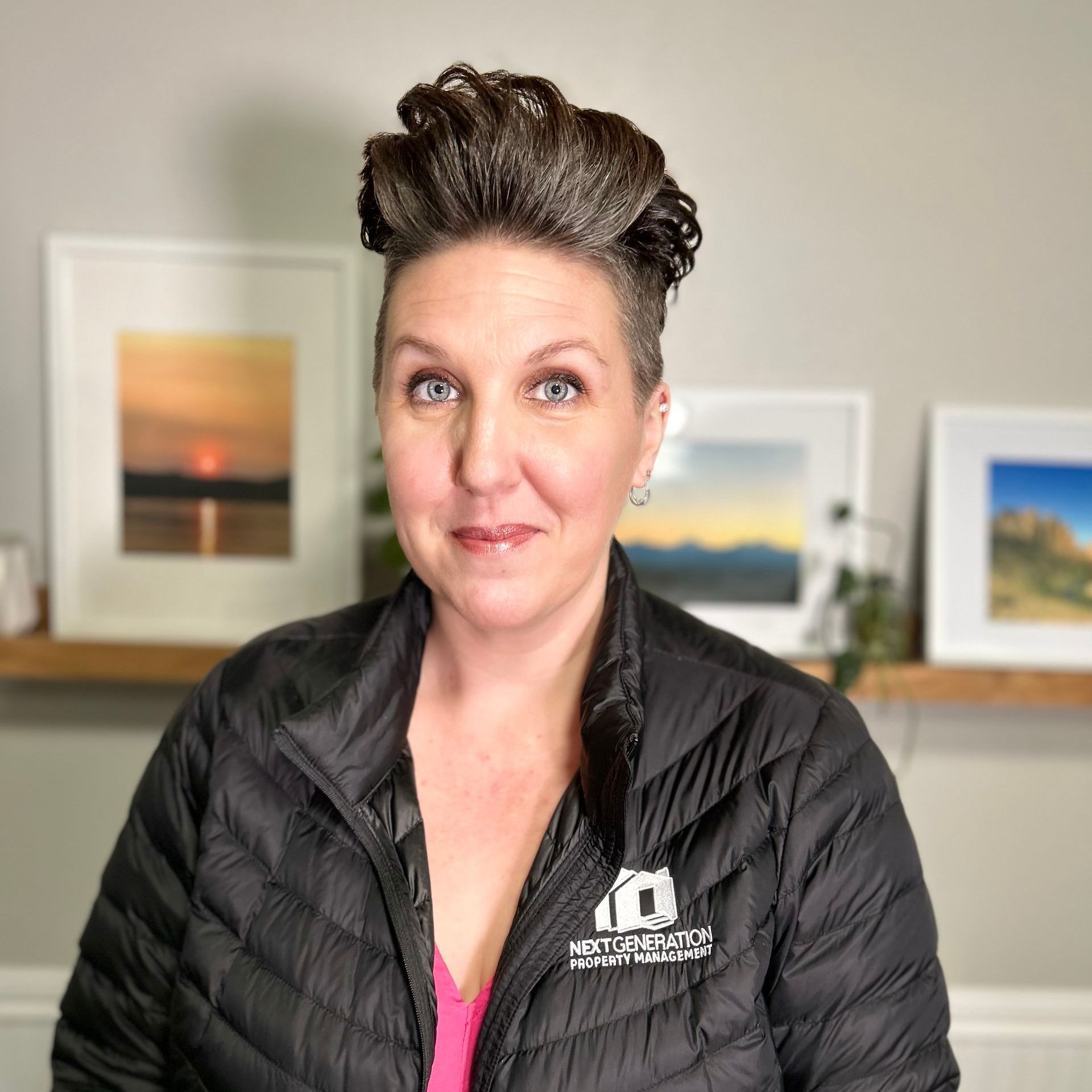 A woman wearing a black jacket is standing in front of a shelf with pictures on it.