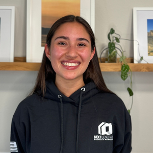 Woman smiling, wearing a black hoodie with logo, standing in front of framed artwork and a shelf with plants.