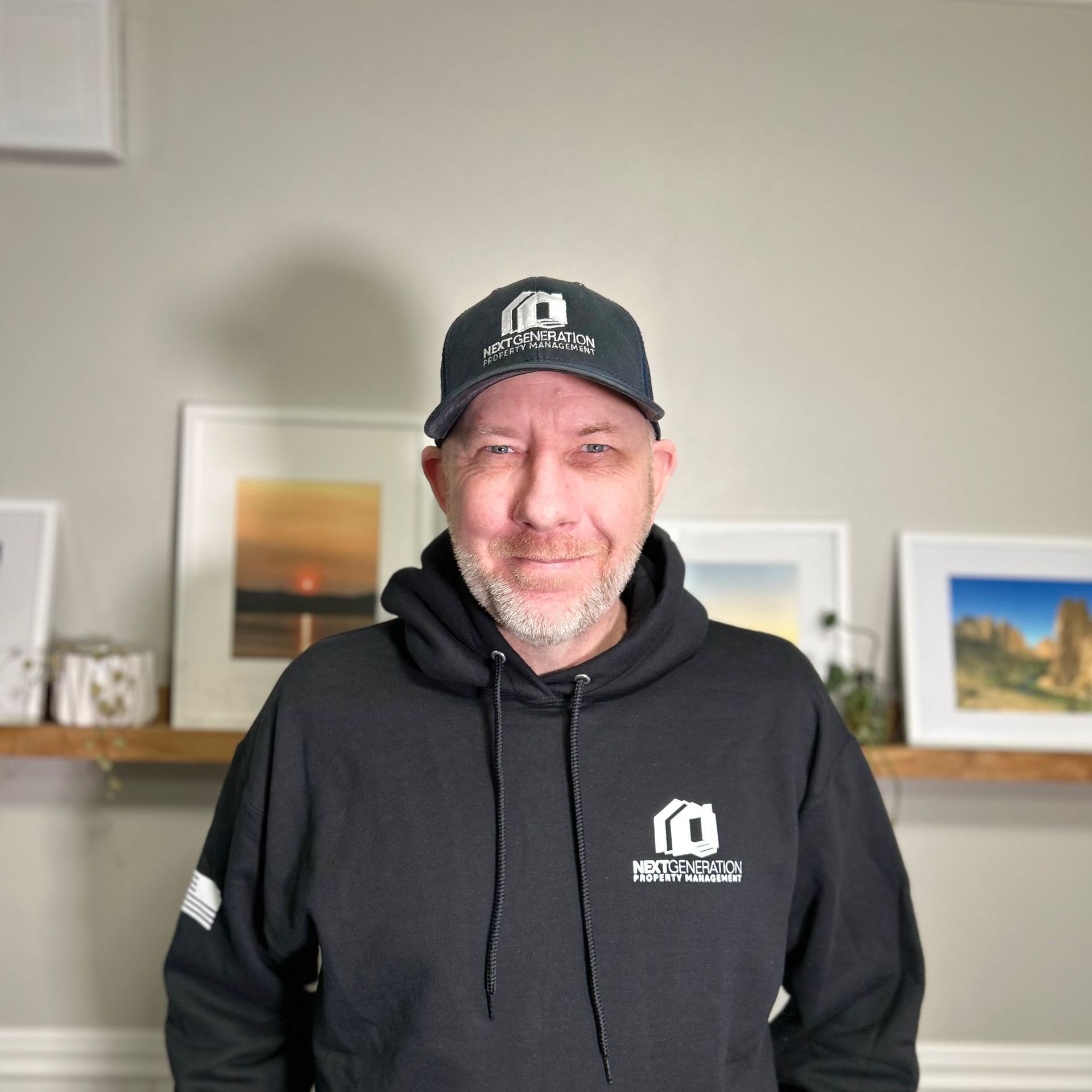 A man wearing a hat and a hoodie is standing in front of a shelf with pictures on it.