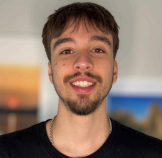 Smiling man with brown hair, goatee, and black shirt. Background features artwork and a blurry wall.