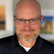 A bald man with a beard and glasses is wearing a black polo shirt.