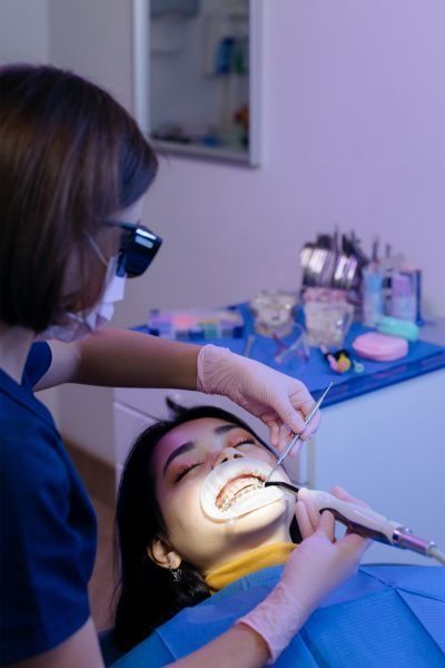 Dentist examining patient's teeth; both wearing masks. In dental chair, under bright light.