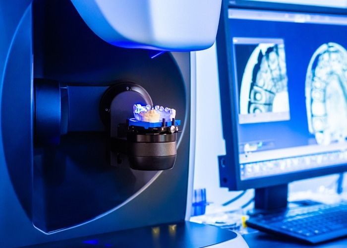 A dental scanner is examining a tooth, with a computer screen displaying an image in a blue-lit lab.