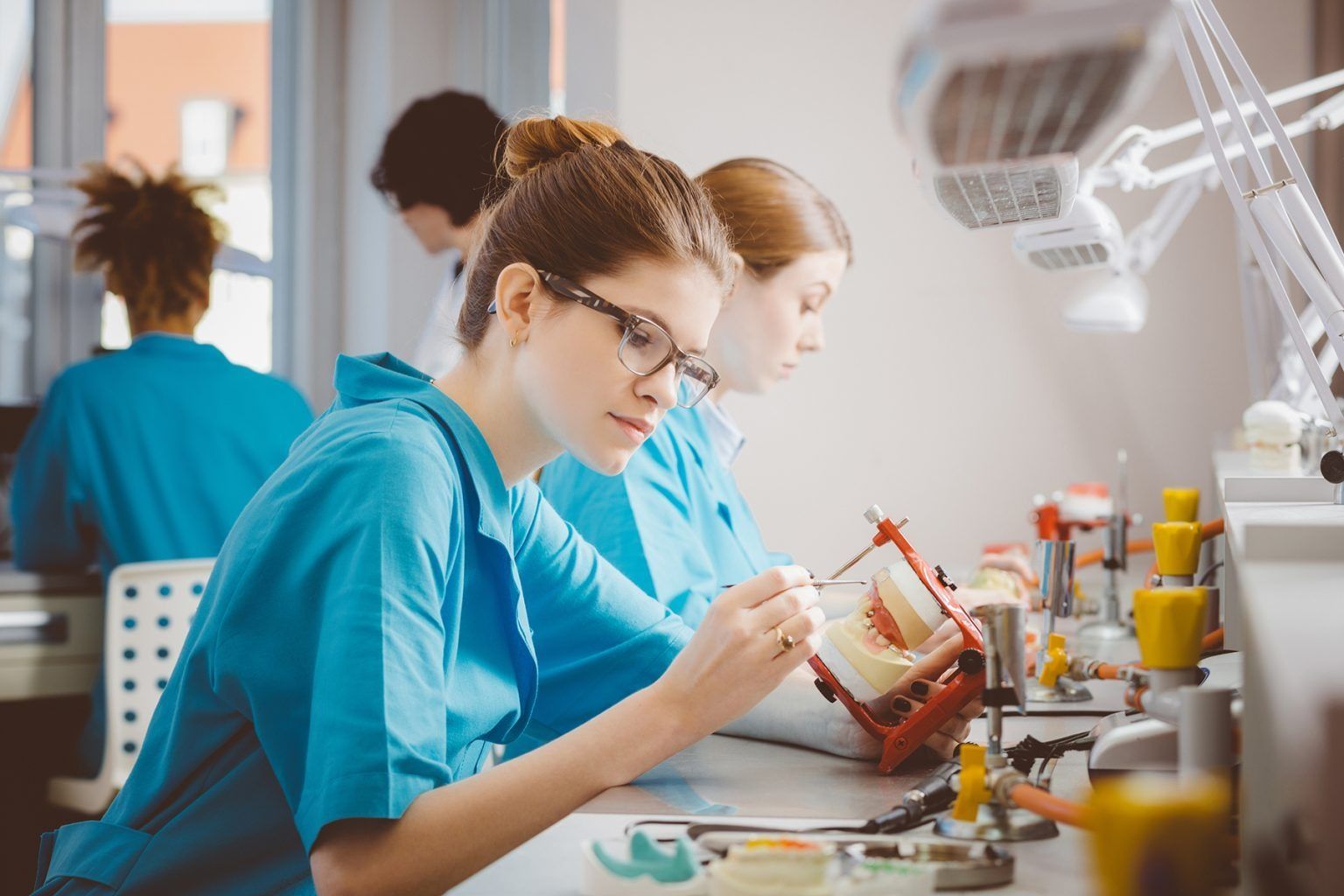 Dental students in blue scrubs working on dental models in a brightly lit lab.