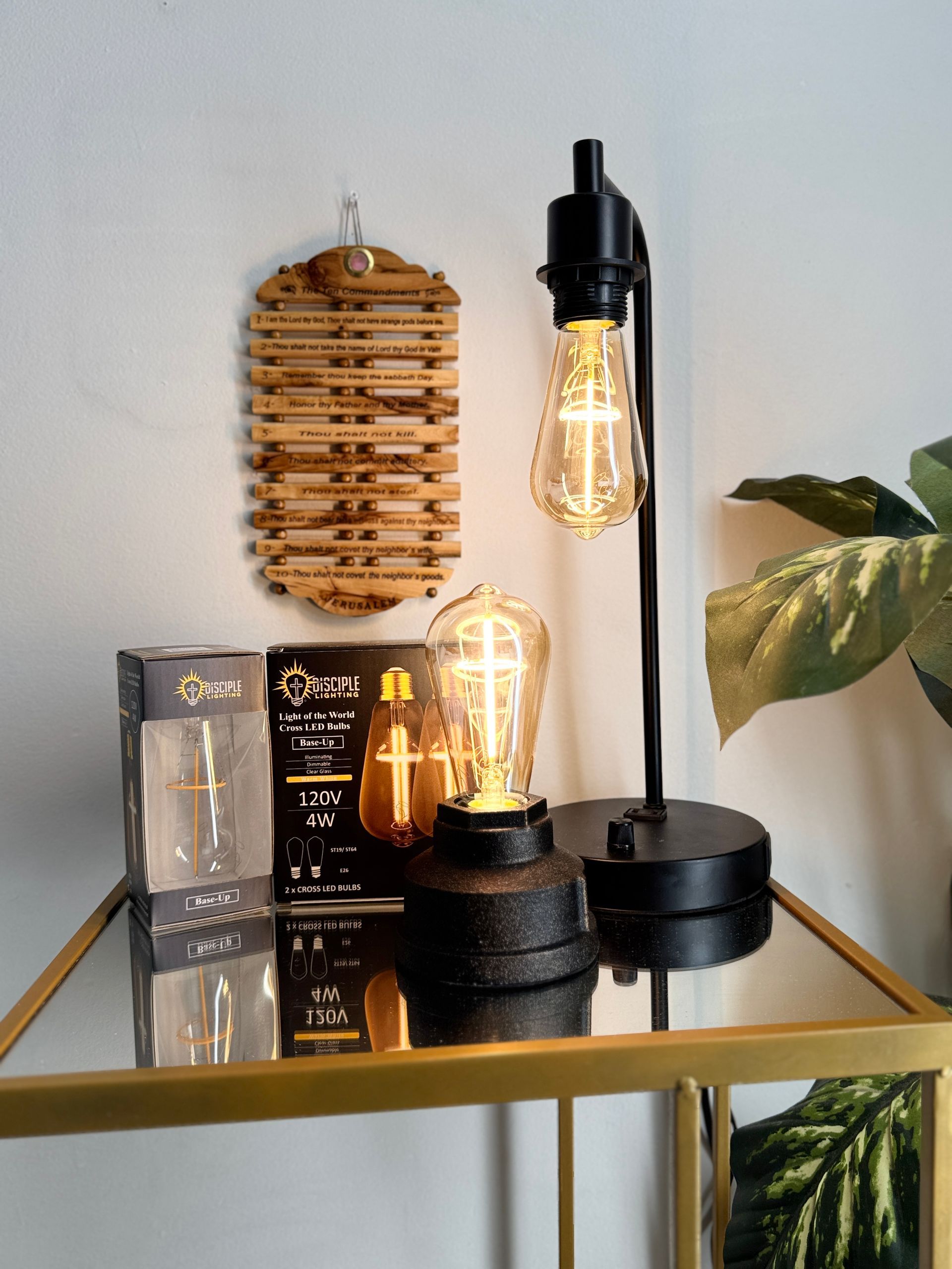 Black desk lamp with Edison bulb on a glass table next to another bulb and box, wooden wall decor.