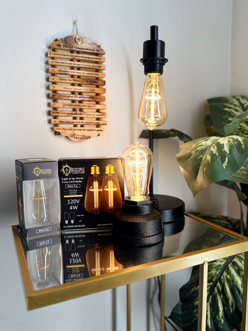 A table lamp with a decorative bulb, next to other decorative items, with a wall hanging on a gold table.
