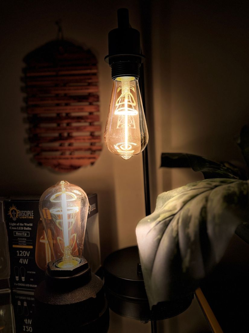 Two lit Edison bulbs, one hanging and one on a base, in a dimly lit room with an artistic wall hanging.