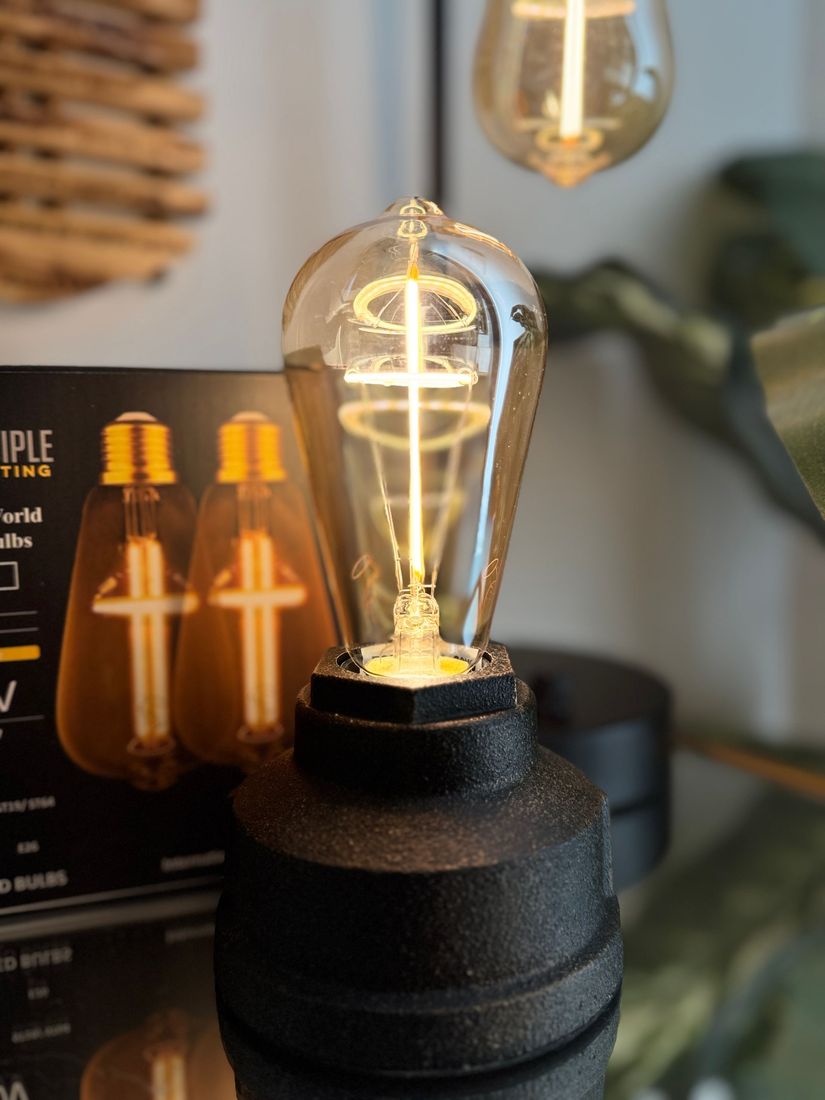 Close-up of a lit Edison-style LED lightbulb on a black base. Box with similar bulbs visible in the background.