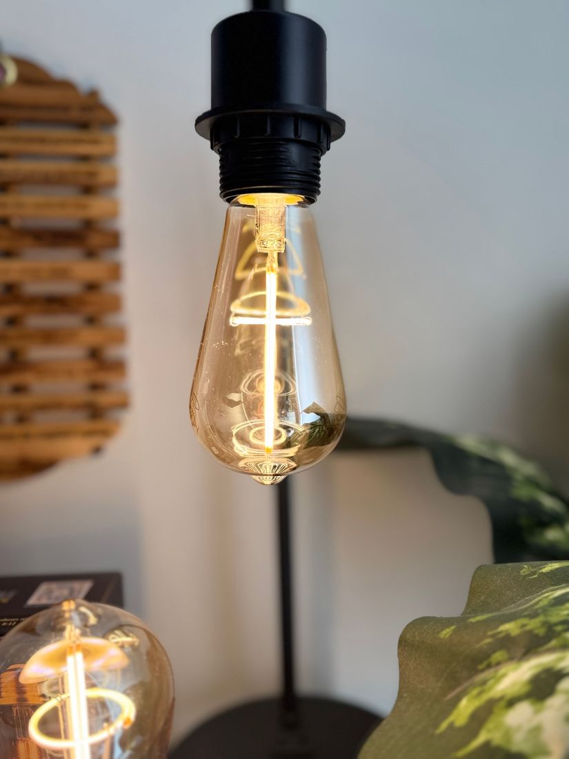 Close-up of a lit, teardrop-shaped Edison bulb in a black socket. Other decor is visible in the background.