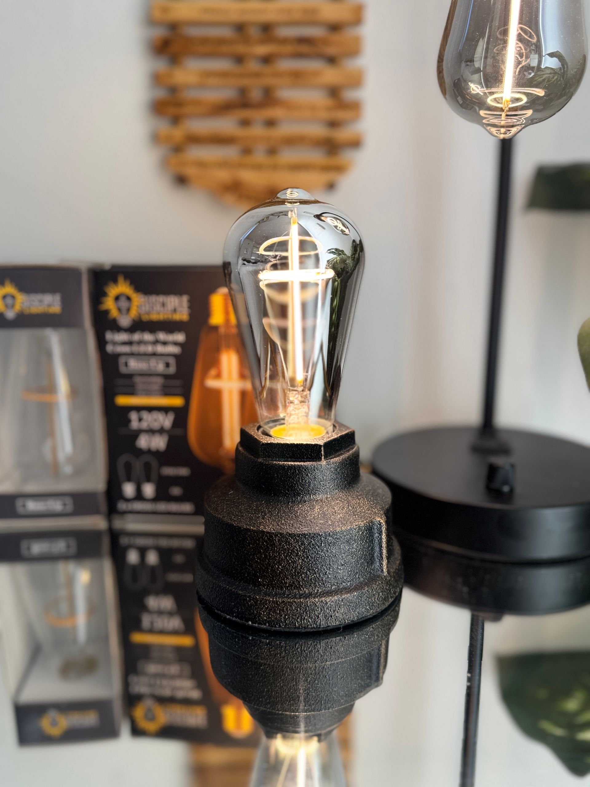 Black and gold lamp with Edison bulb, set on a reflective surface. Product packaging in the background.