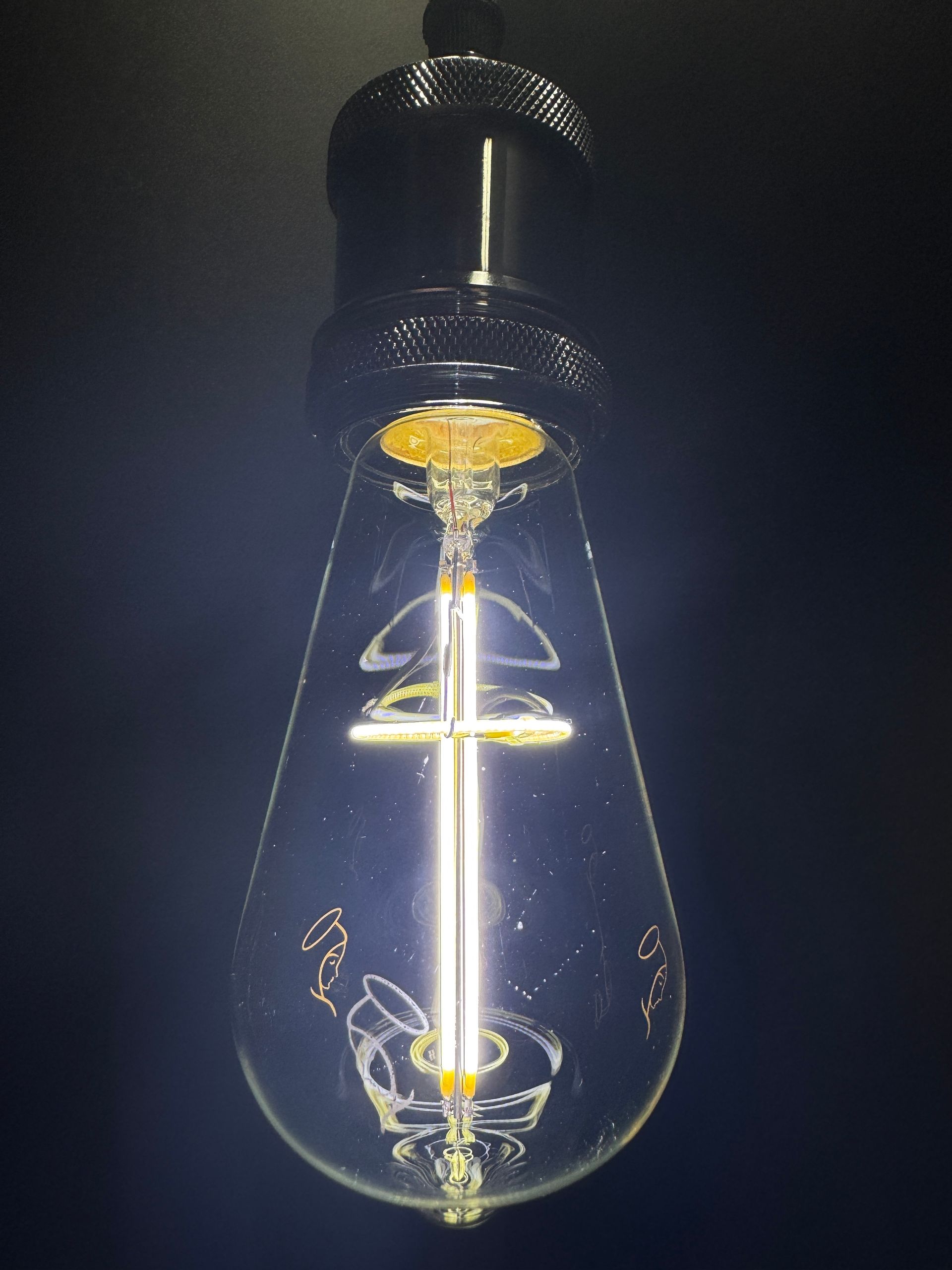 Lit, teardrop-shaped Edison bulb with visible filament and internal supports, against a dark background.