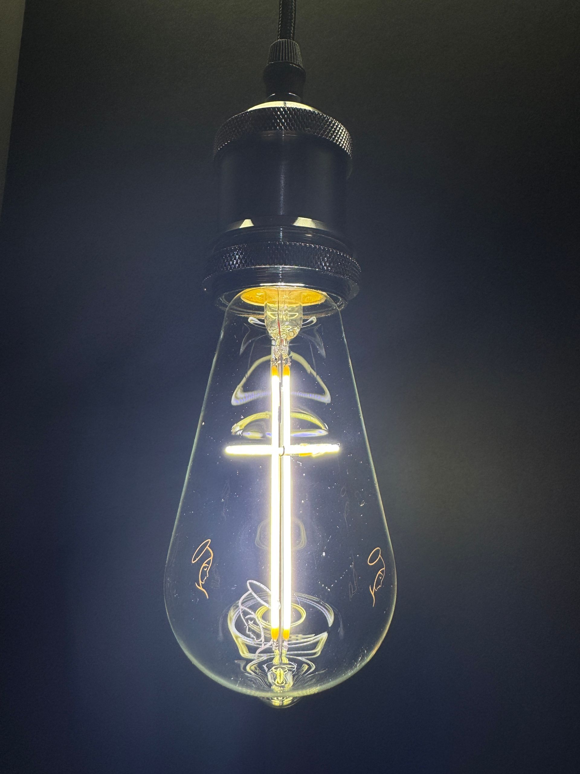 Lit, teardrop-shaped Edison bulb hangs from a black cord, glowing with a vertical filament.