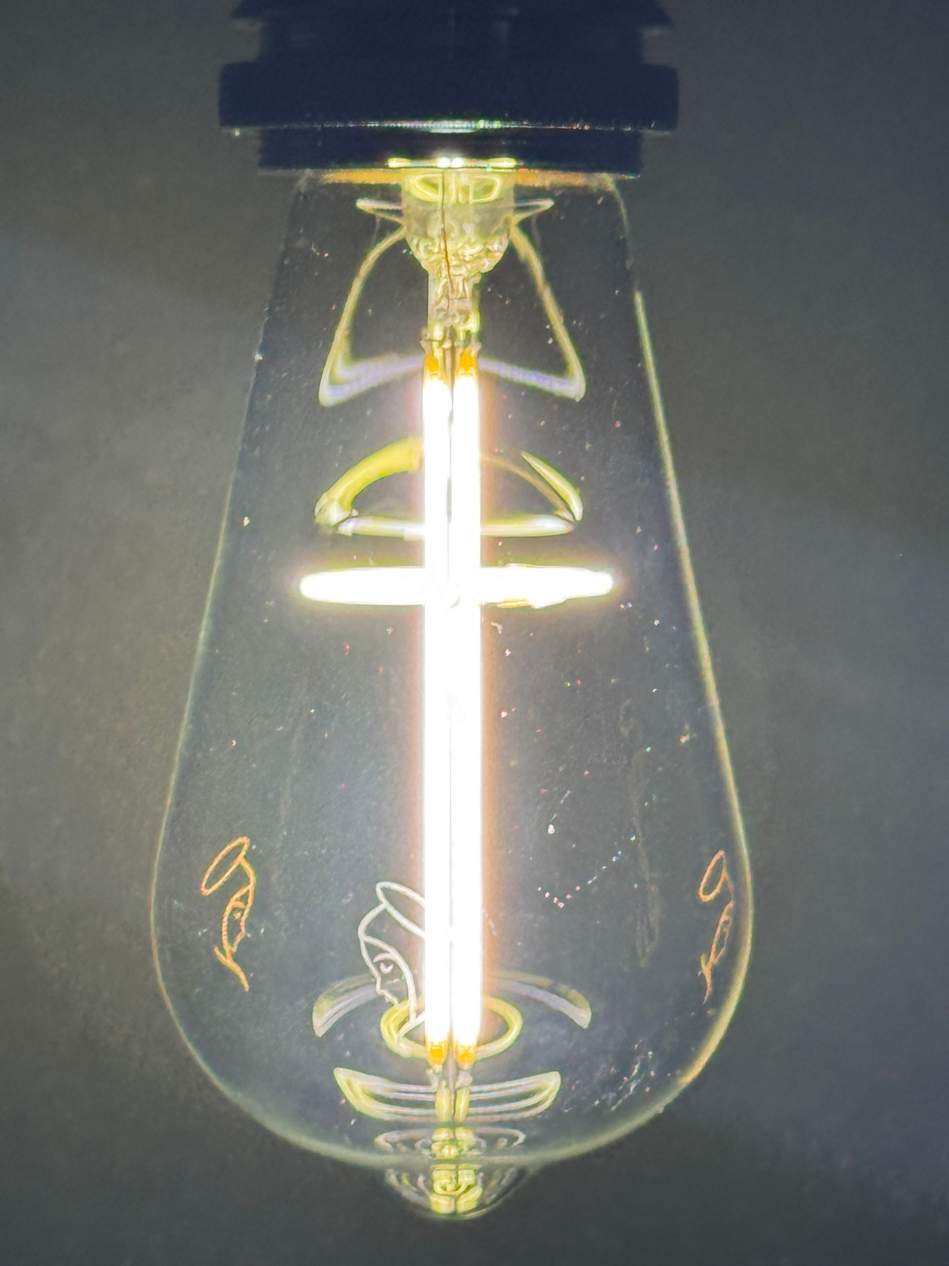 Lit teardrop-shaped Edison lightbulb with vertical filament and decorative internal elements, emitting warm light.