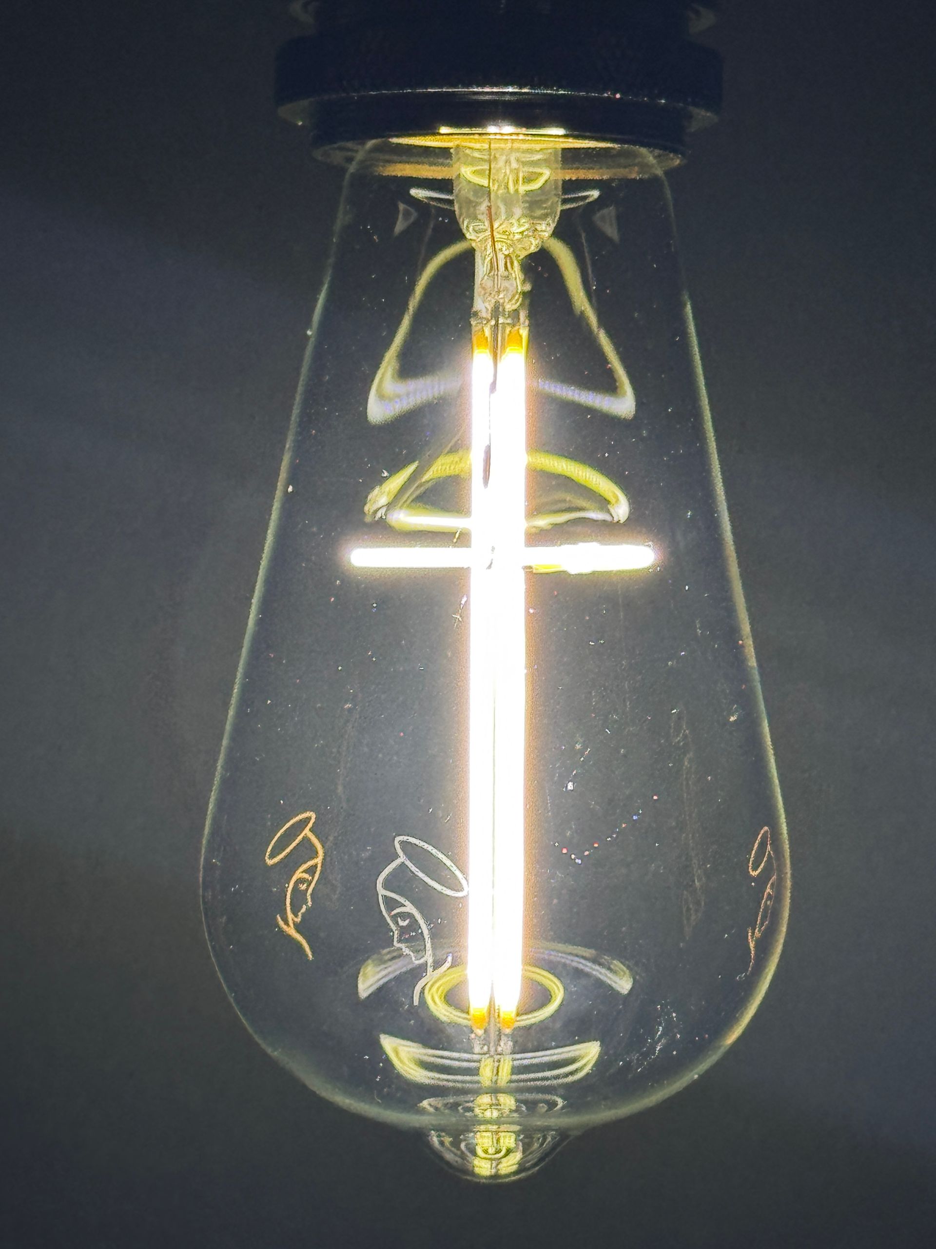 Lit LED lightbulb with cross filament and etched religious imagery.