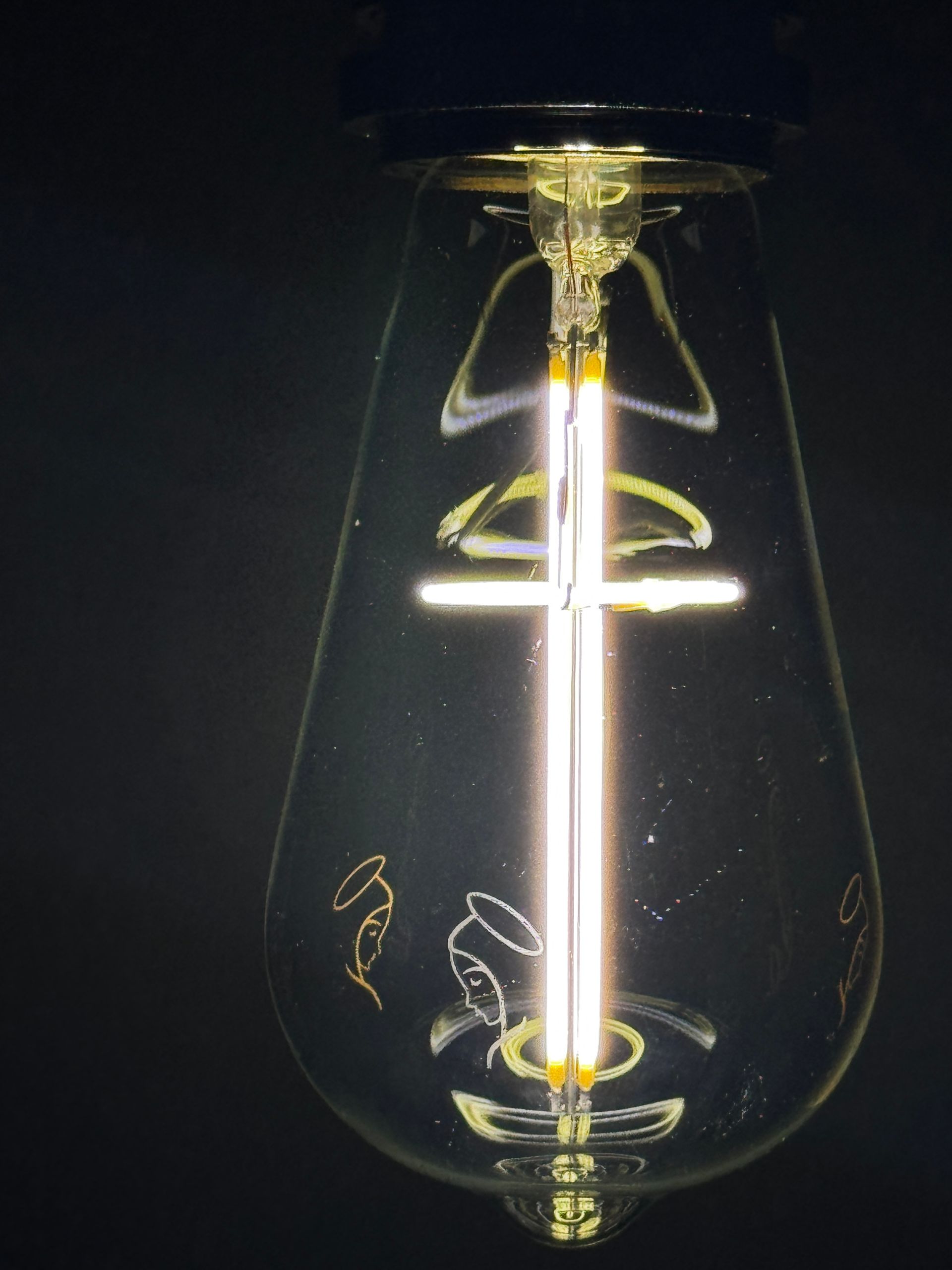 Lit teardrop-shaped light bulb with LED filaments; etched angels on the glass, illuminated against a dark background.