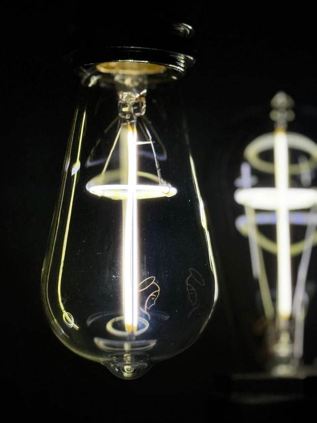 Close-up of lit, teardrop-shaped light bulb with a cross-shaped inner element, black background.
