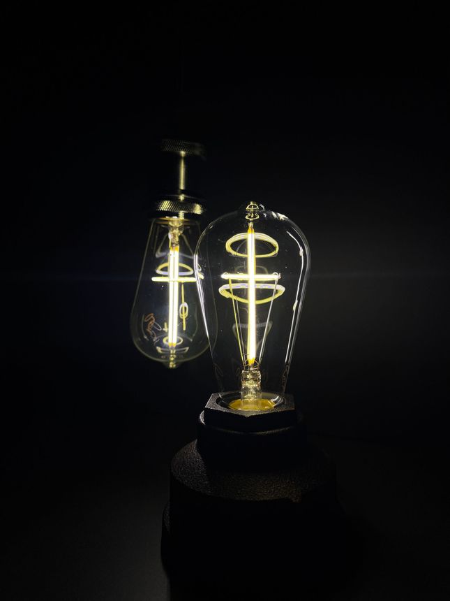 Two ornate light bulbs shaped like teardrops, illuminated with a cross design, against a dark background.