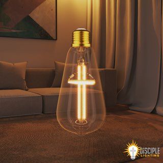 Large lightbulb-shaped lamp with a cross design, indoors on a rug, near a couch. Golden light illuminates the scene.