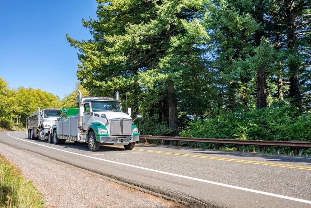 Two Semi Trucks Are Driving Down a Highway Next to Trees — Bulahdelah Towing In Karuah, NSW