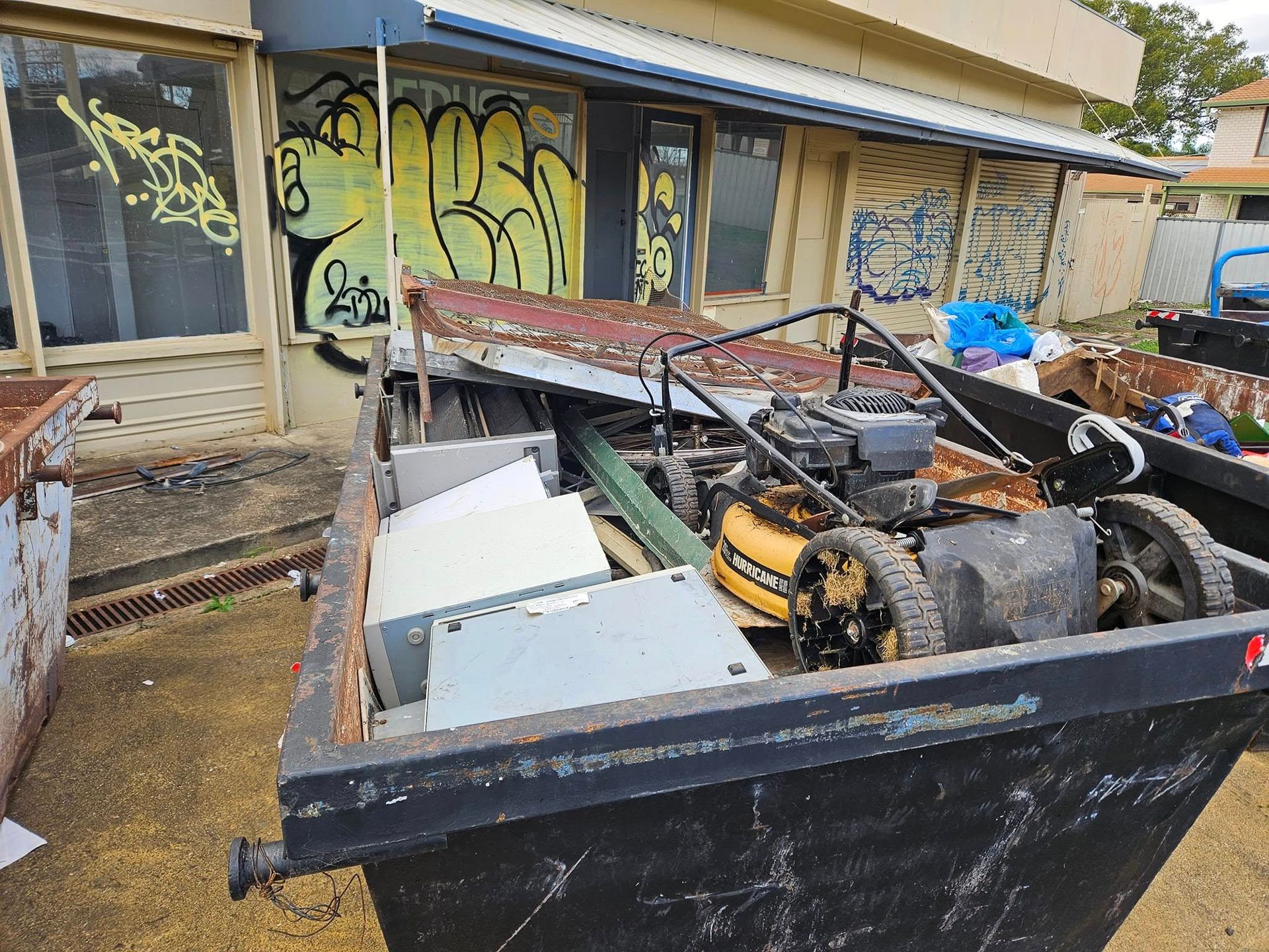Dumpster filled with junk in front of a graffiti-covered building. Includes a lawnmower, metal scraps. - Tamworth Skips and Rubbish Removal in South Tamworth, NSW