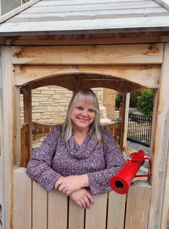 A smiling person in a purple patterned top leans on a wooden structure featuring a red toy telescope.