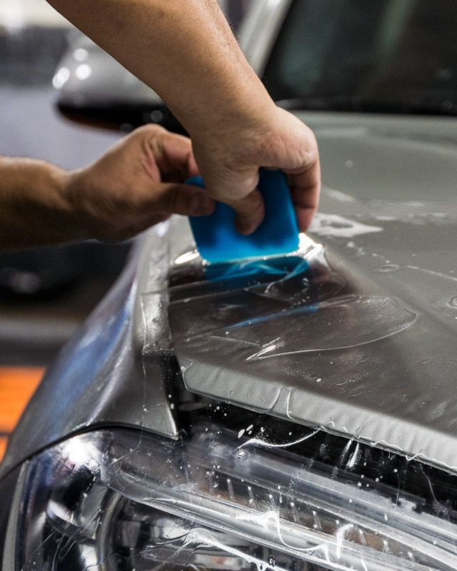 A person is using a blue squeegee to remove plastic from the hood of a car