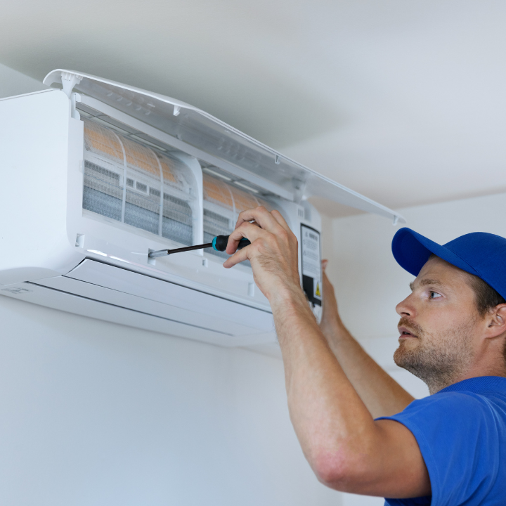 A man in a blue hat is fixing an air conditioner with a screwdriver