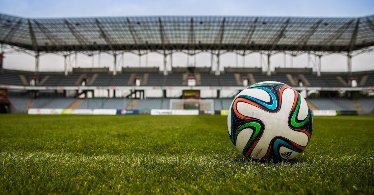 A soccer ball is sitting on top of a soccer field in front of a stadium.