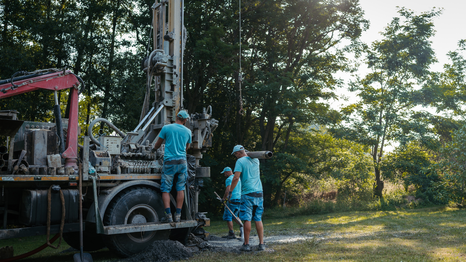 Three workers in matching blue shirts operate a truck-mounted drilling rig in a sunny, wooded outdoor area.