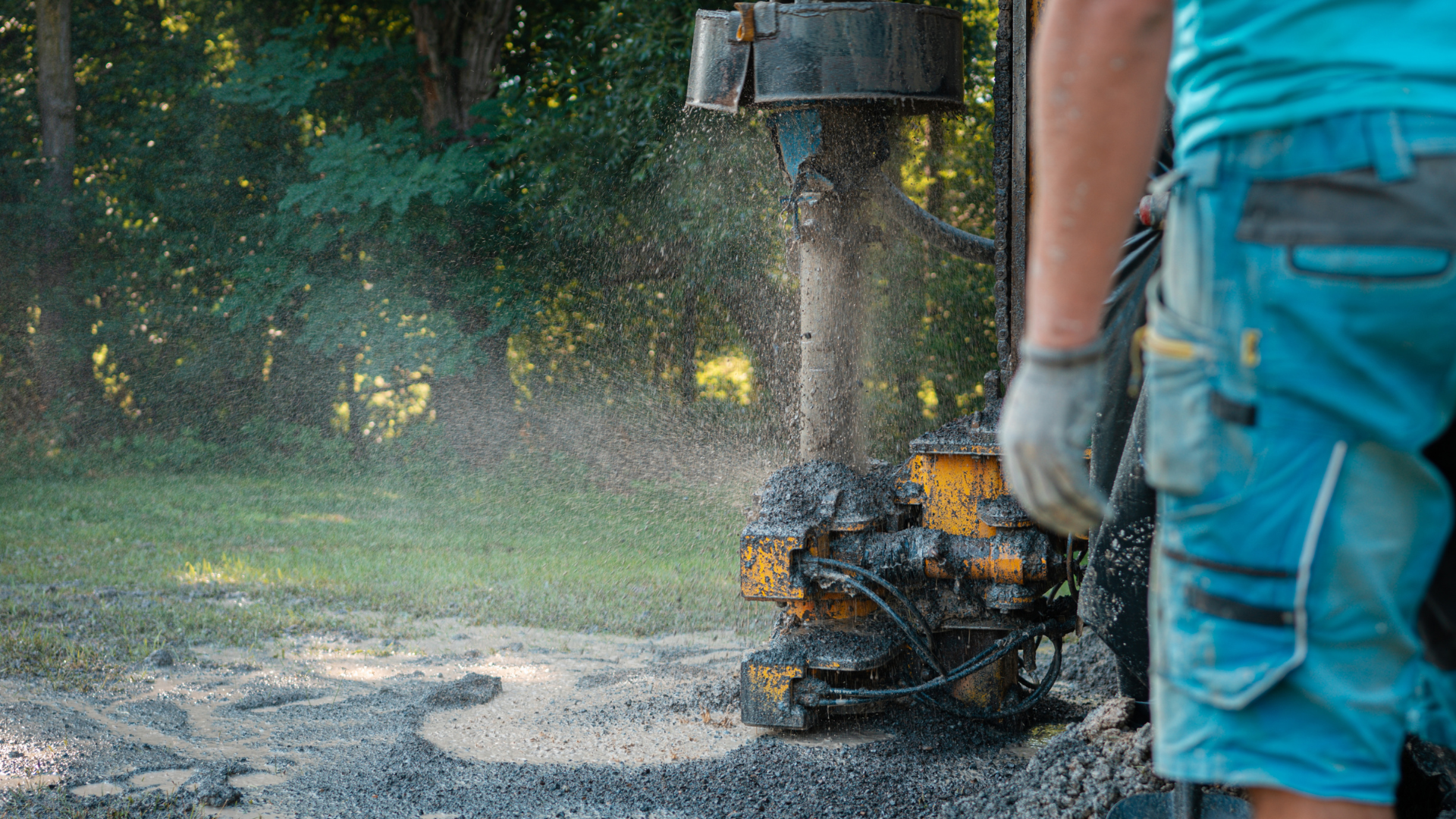 A worker in blue workwear stands beside a yellow industrial drill boring into the ground outdoors.